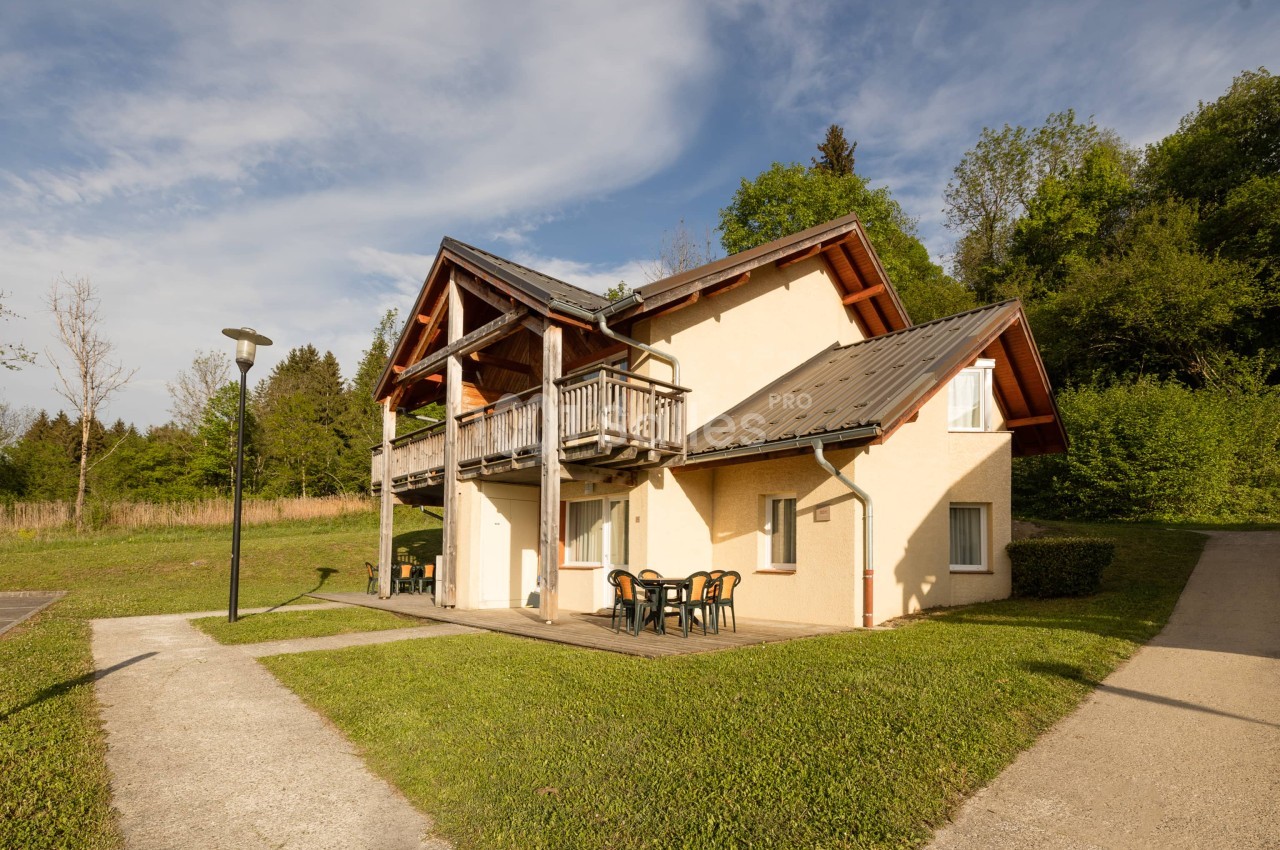 Maison de plain-pied et étage en crépi beige à toit métallique brun, avec balcon en bois et terrasse dallée meublée de tables et chaises de jardin, entourée de pelouse et d'arbres. Let me check length: roughly 170 characters, too long. Need ≤125. Maison