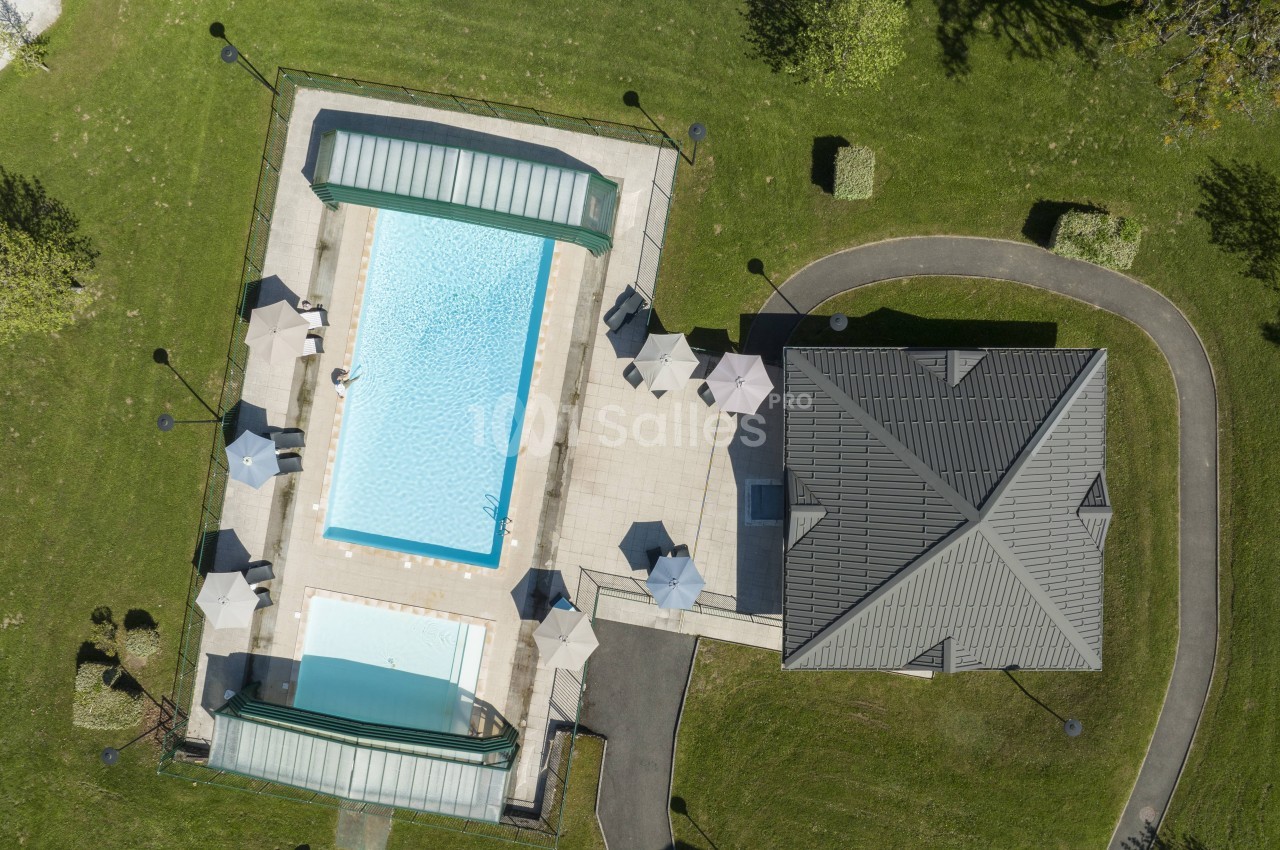 Vue aérienne d'une grande piscine rectangulaire et d'une petite piscine, bordées de parasols et de transats, à côté d'un bâtiment au toit gris. Wait, I must not begin with "Vue de". The rule forbids starting with "Photo de", "Image de", "Vue de". "Vue