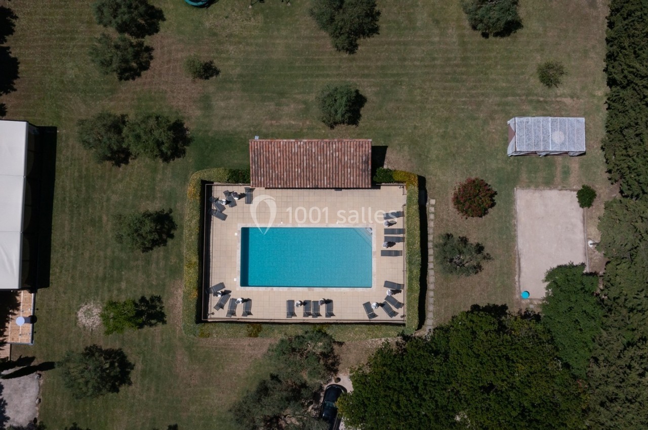 Vue aérienne d'une piscine rectangulaire entourée de chaises longues, située dans un jardin arboré.