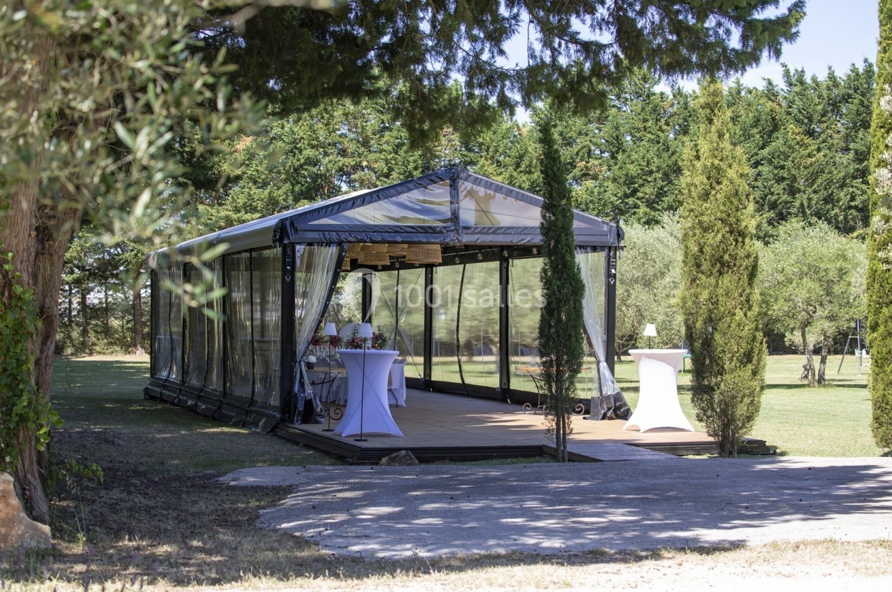 Structure en verre et métal installée dans un jardin, aménagée avec des tables et des chaises pour un événement.