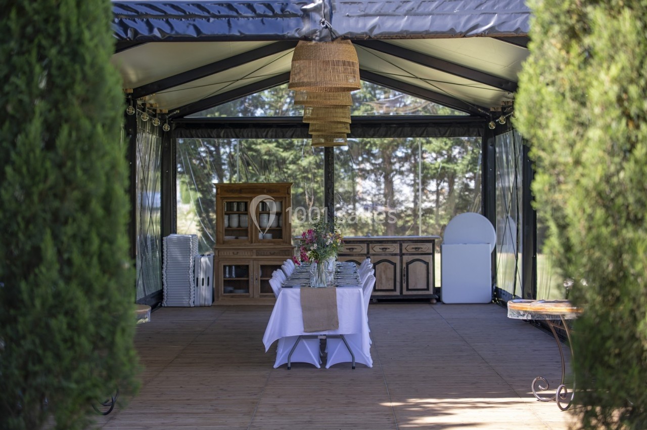 Intérieur d'une tente aménagée avec une table dressée, des meubles en bois et des lampes suspendues, entourée de verdure.