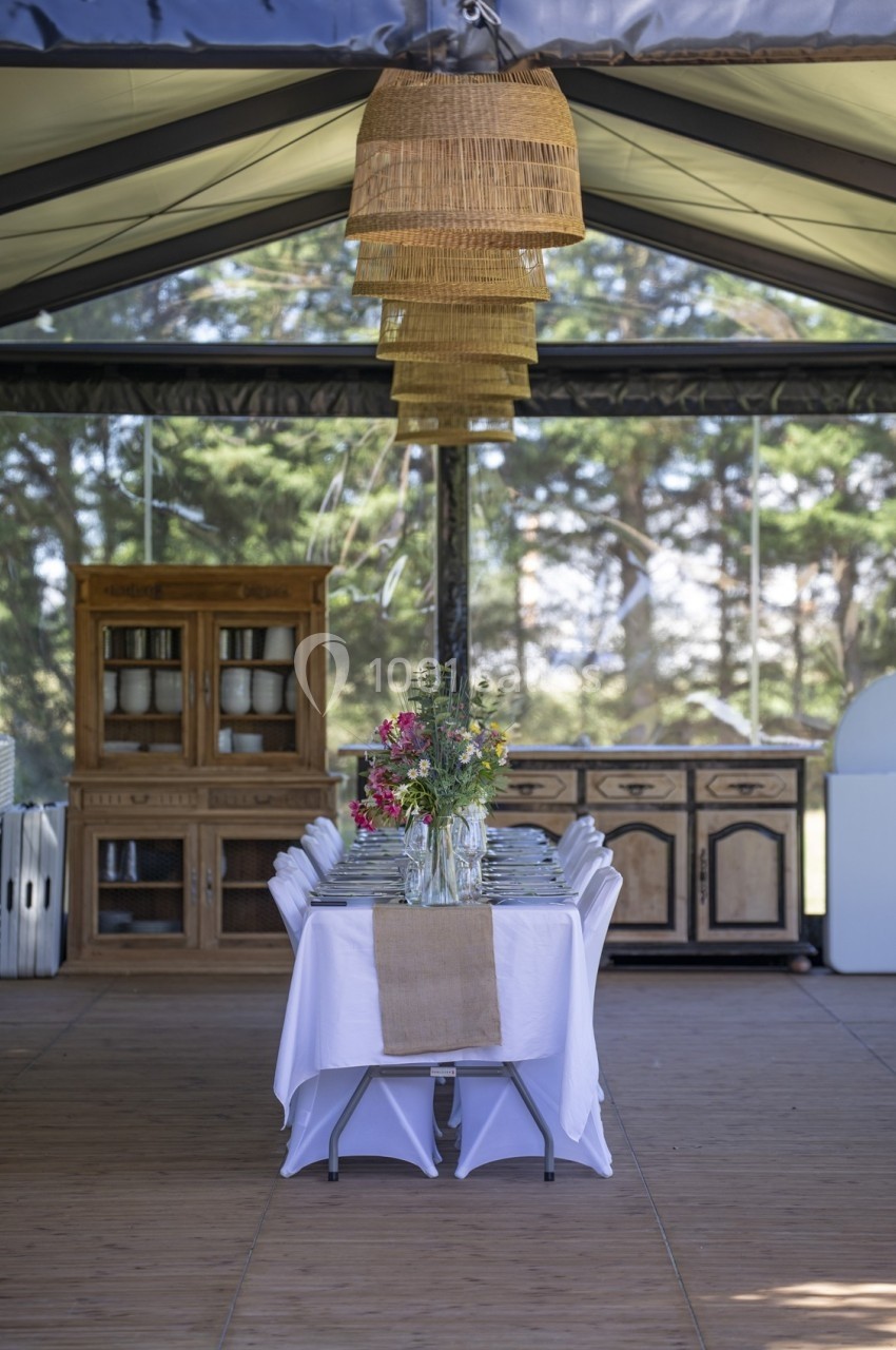 Table dressée avec nappe blanche, chemin de table en toile de jute et vase de fleurs, sous un abri avec meubles en bois.