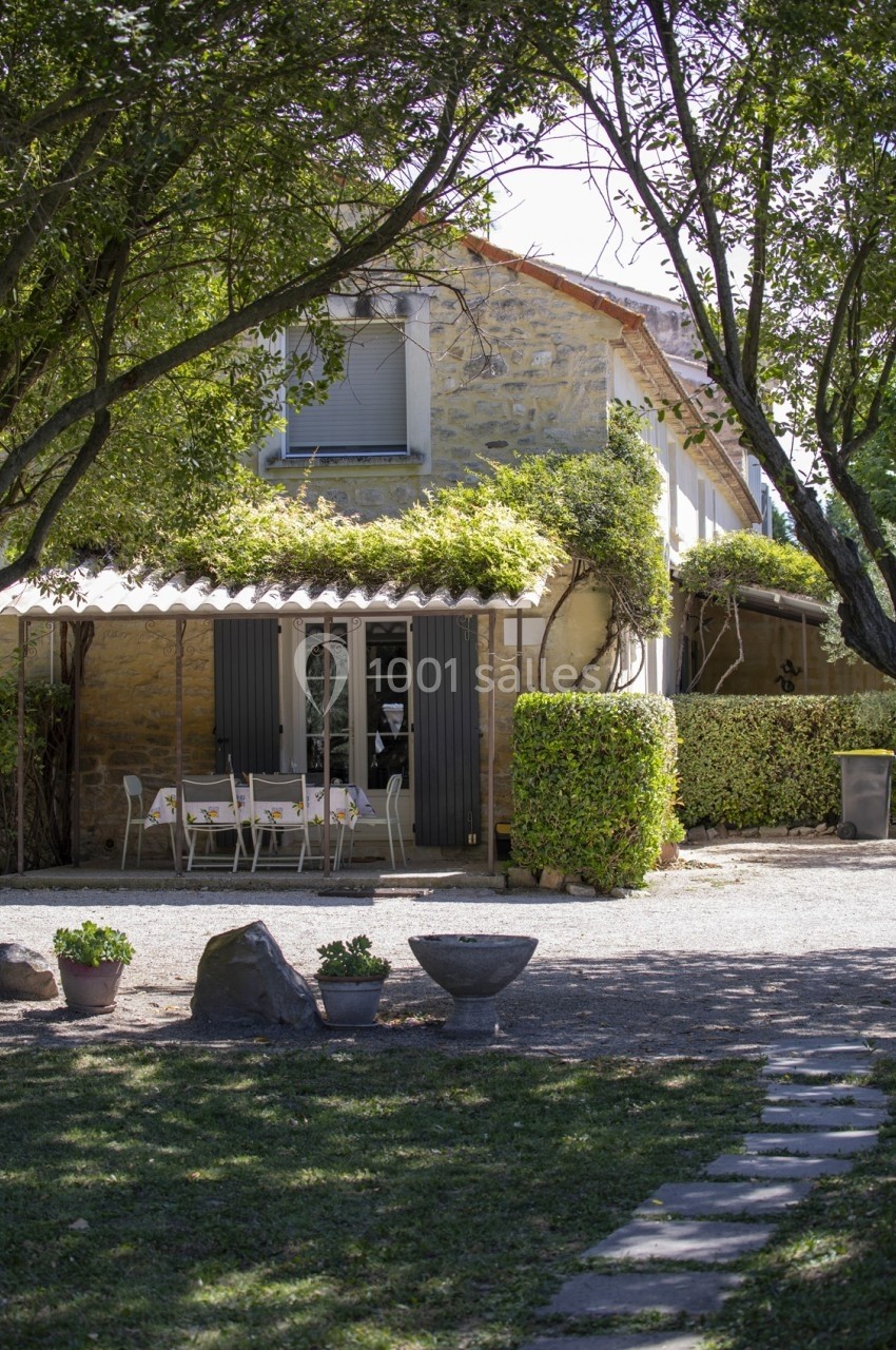Maison en pierre avec terrasse ombragée, table dressée, entourée de végétation et d'un jardin avec allée en pierre.