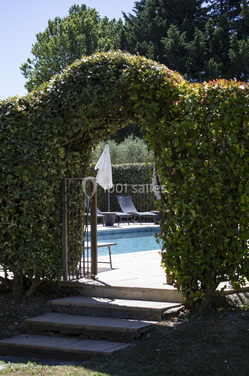 Arche végétale donnant sur une terrasse avec piscine, transats et parasol, entourée de haies et d'arbres.