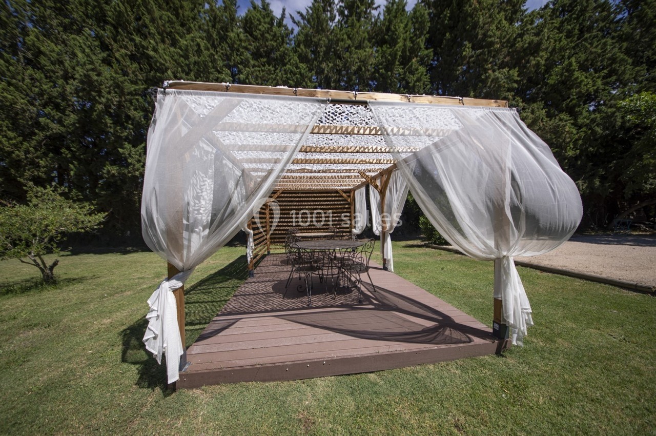 Pergola en bois avec rideaux blancs translucides, installée sur une terrasse en bois dans un jardin verdoyant.
