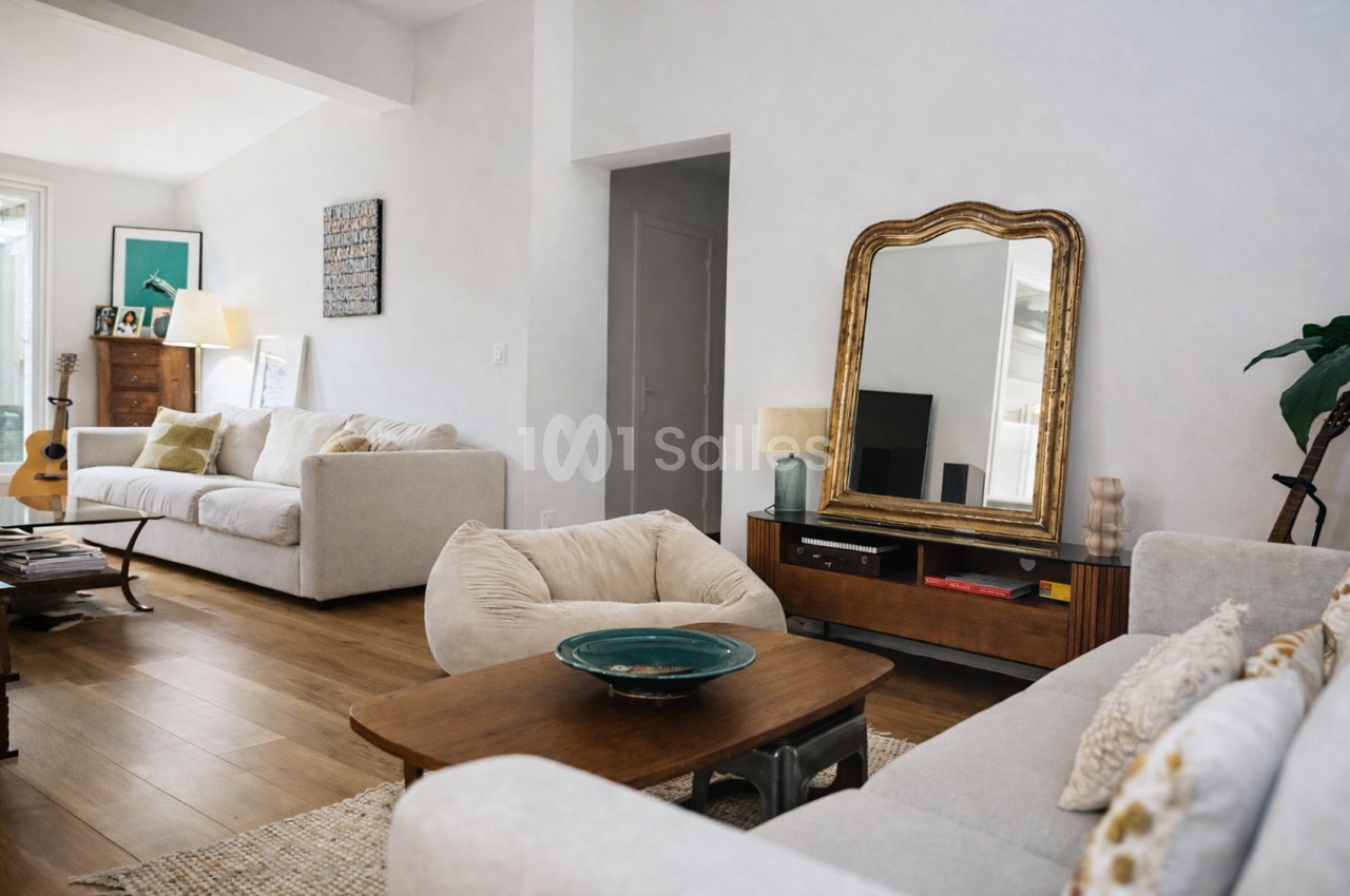 Salon spacieux aux murs blancs avec canapés beige clair, table basse en bois et miroir doré, sol en parquet.