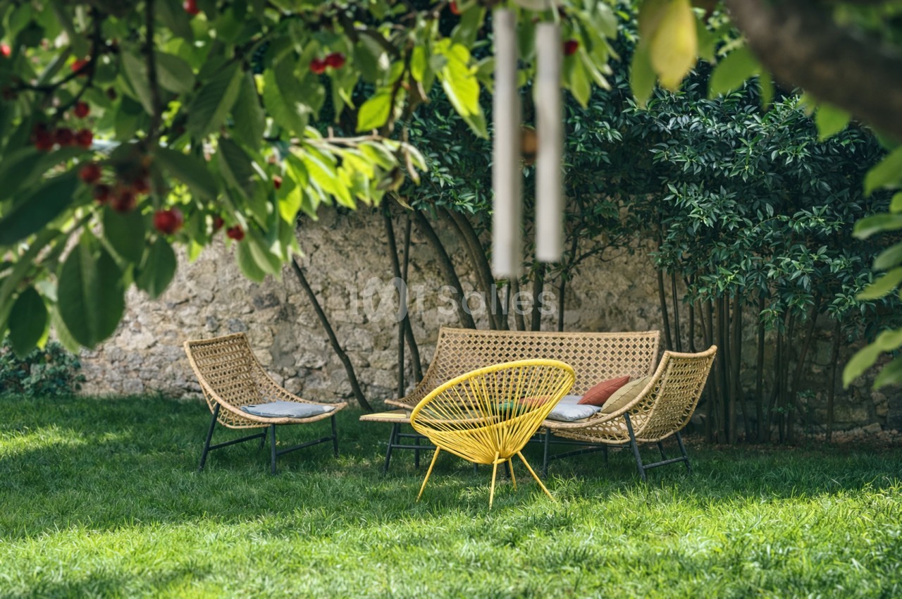 Jardin ombragé avec trois fauteuils en rotin face à une table basse jaune, mur en pierre et végétation.