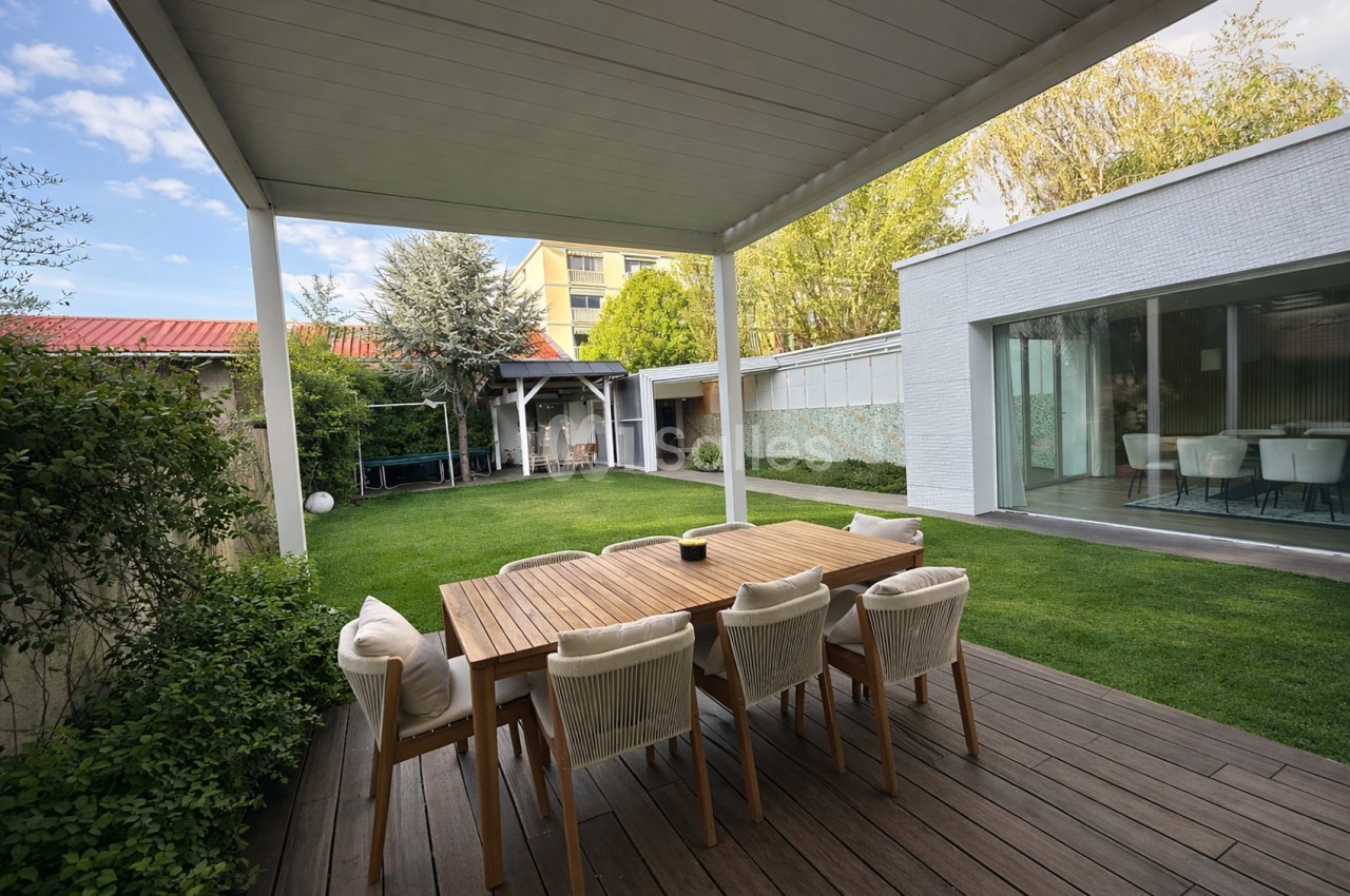 Terrasse couverte avec table en bois, chaises beiges et jardin paysager en arrière-plan.