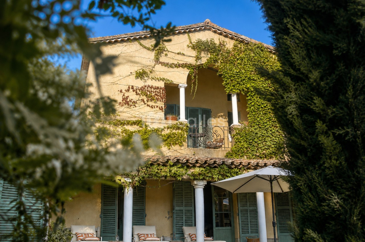 Façade ocre de villa provençale avec colonnes blanches, volets verts, lierre grimpant, terrasse couverte et parasol au rez-de-chaussée.