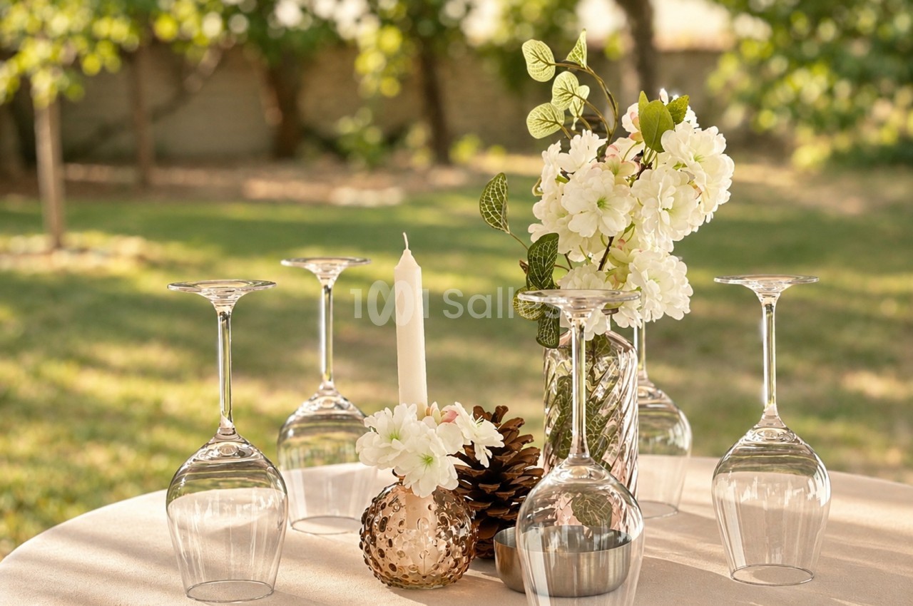 Centre de table avec fleurs blanches en vase, verres vides et bougie, jardin en arrière-plan.
