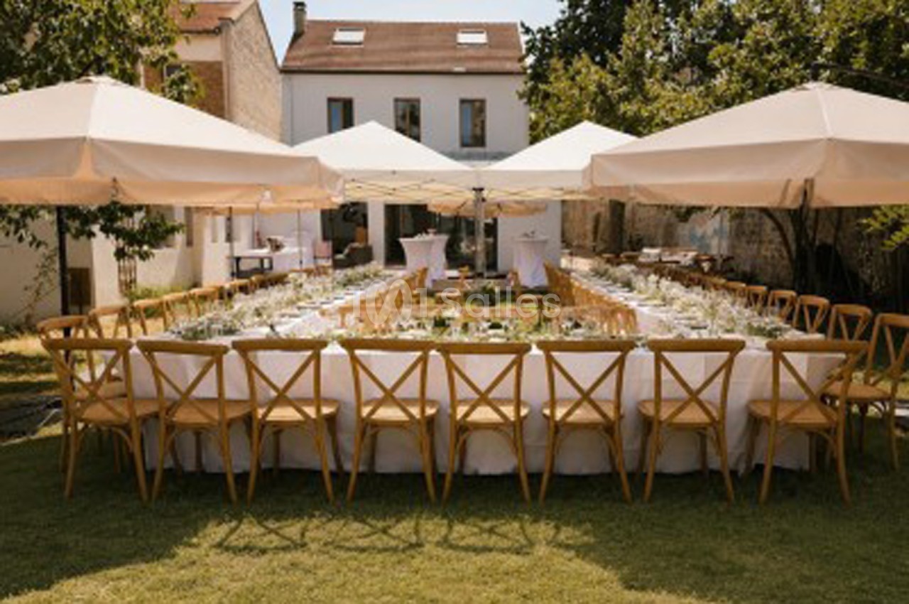 Table dressée en U sous voiles beige dans jardin, chaises bois, bâtisse pierre en arrière-plan.