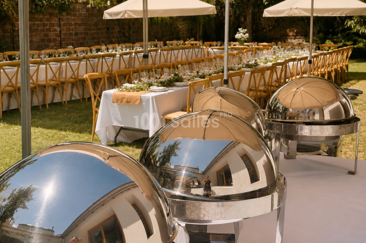 Chafing dishes en inox dans jardin avec tables dressées sous parasols blancs et chaises en bois.