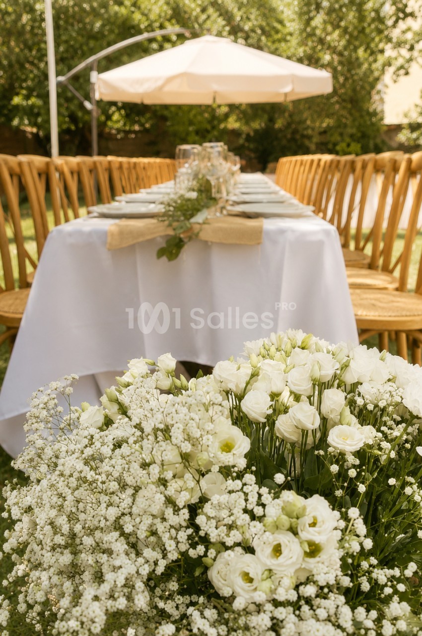 Jardin avec table ronde dressée sous parasol blanc, chaises jaunes et compositions florales blanches en premier plan.