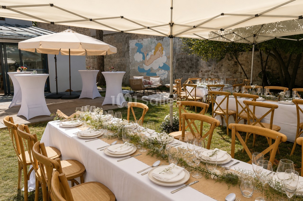 Terrasse couverte dressée pour un événement avec tables longues blanches, chaises en bois, parasol blanc et tonnelles de toile crème.