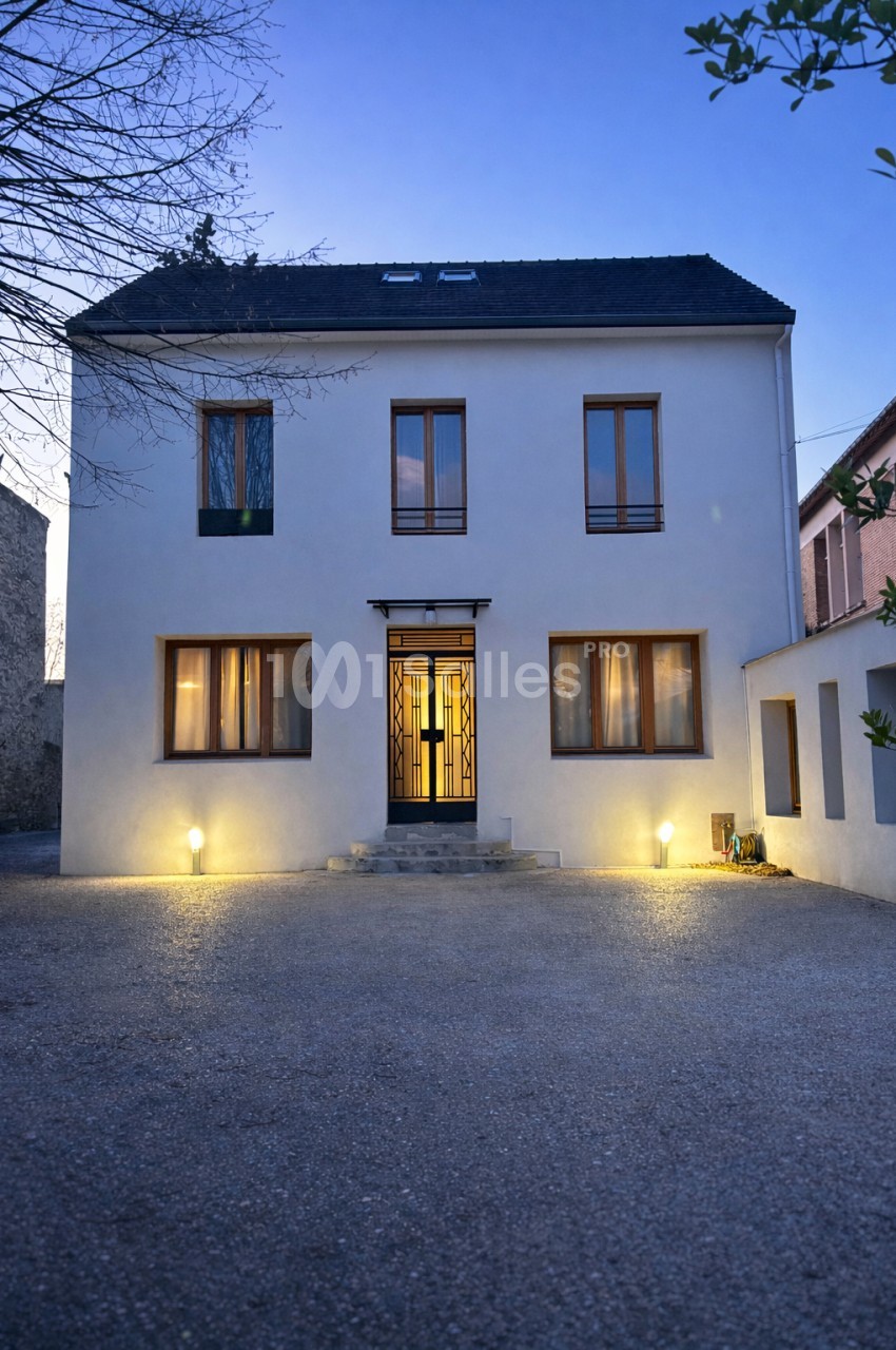 Façade blanche de maison contemporaine illuminée le soir, entrée centrale et cour pavée.