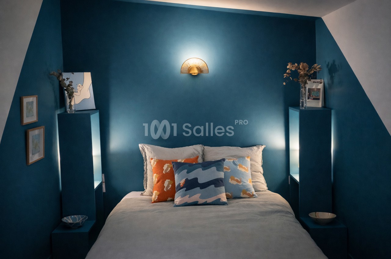 Chambre aux murs bleu canard avec lit double, coussins colorés, deux chevets intégrés et applique murale dorée.