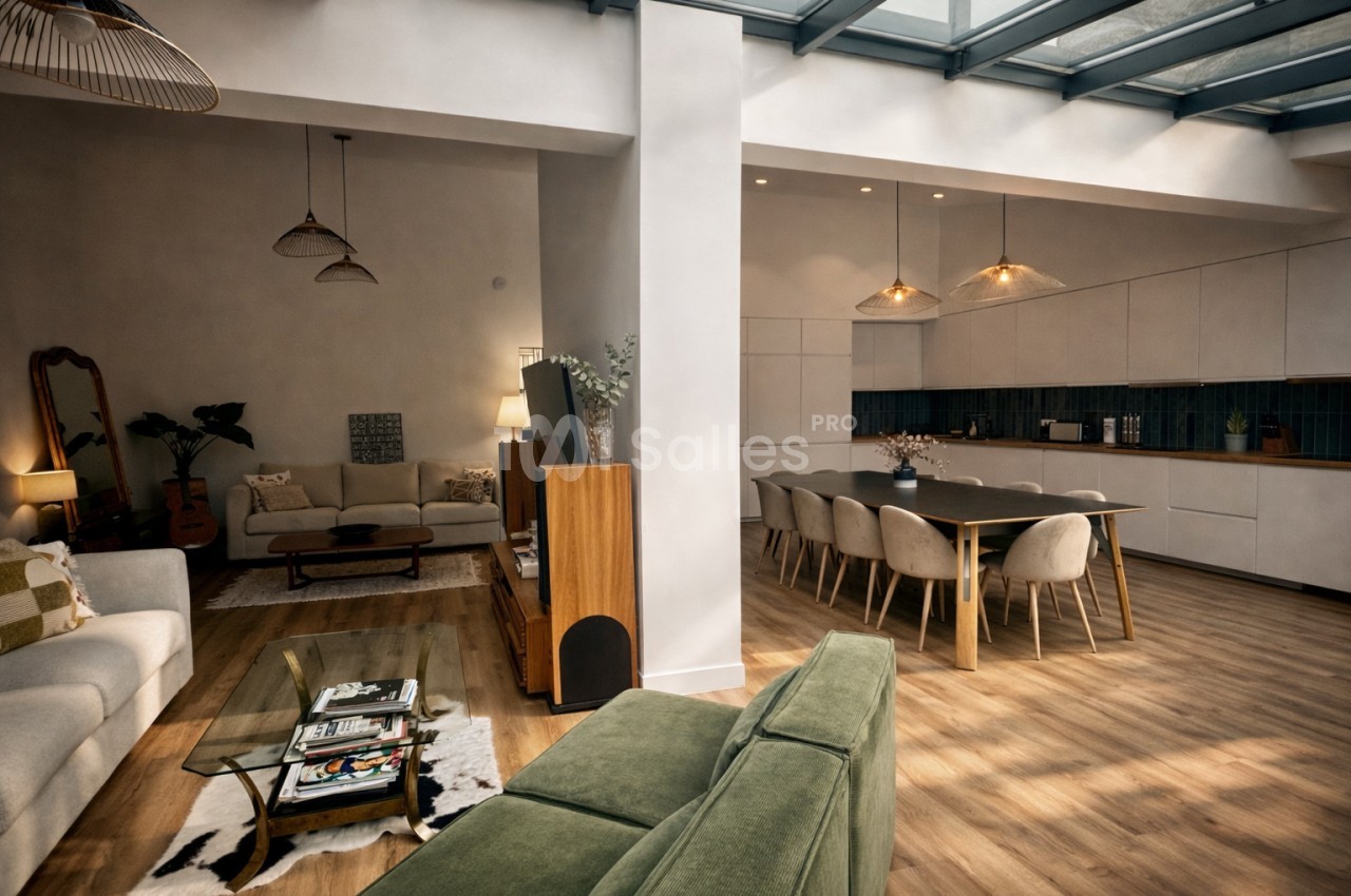 Intérieur d'un loft aux volumes ouverts - salon avec canapés beige, coin cuisine équipé d'une table et chaises grises, lumière naturelle du plafond.