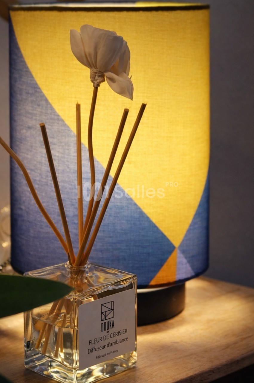 Diffuseur de parfum avec fleur en pétales blancs et tiges dorées devant une lampe cylindrique colorée.