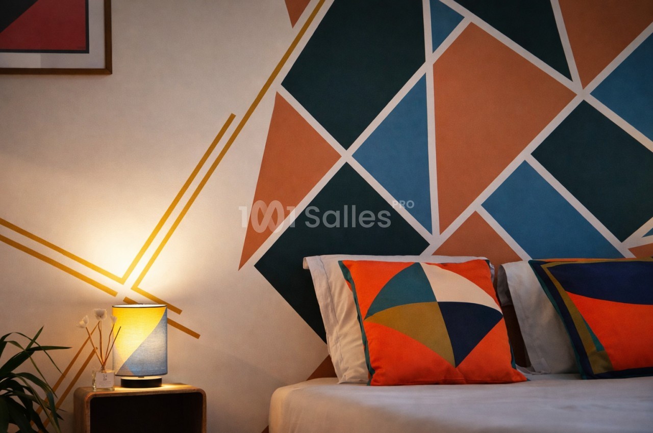 Chambre avec lit blanc, coussins géométriques colorés et mur orné de triangles multicolores - bleu, orange, noir.