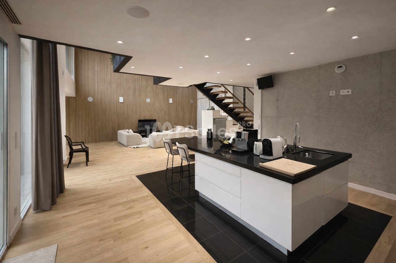Espace ouvert avec cuisine îlot blanche, salon avec canapés beige et escalier bois, sol en parquet clair.
