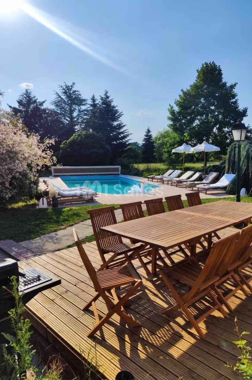 Terrasse en bois avec table et chaises face à une piscine entourée de chaises longues et parasols, arbres en arrière-plan.