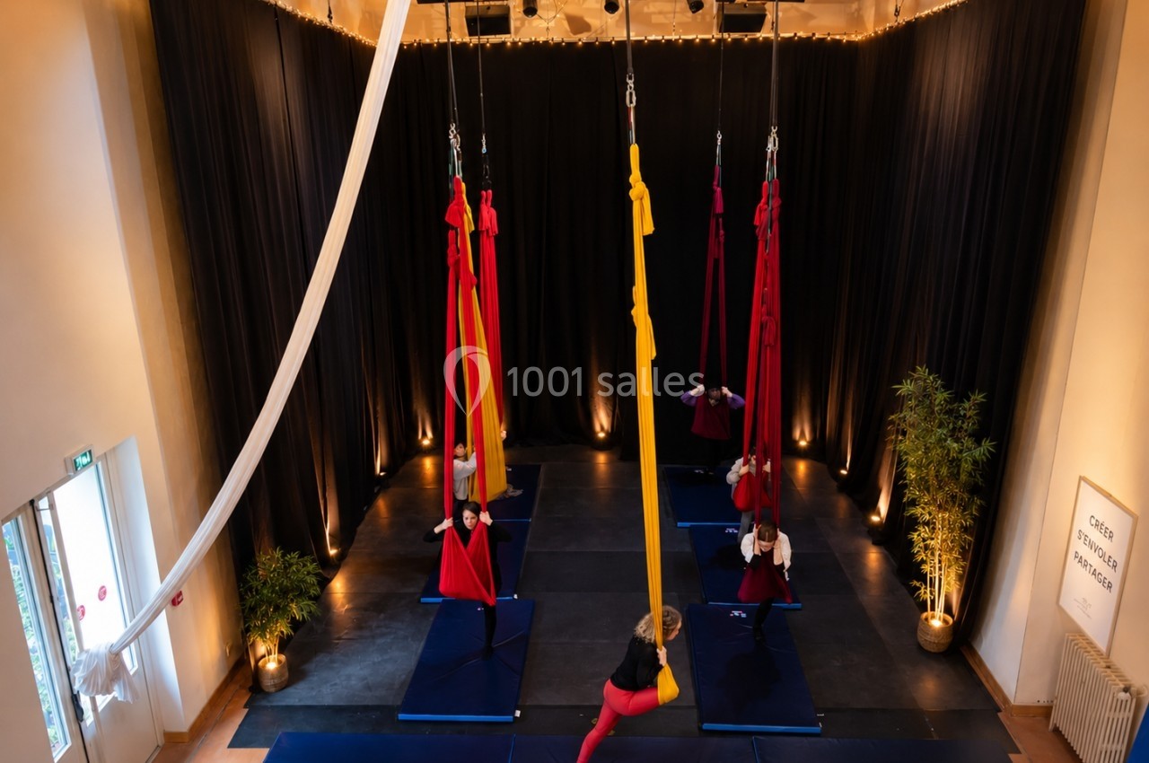 Personnes pratiquant le yoga aérien dans une salle avec des tissus suspendus, éclairage chaleureux et tapis au sol.