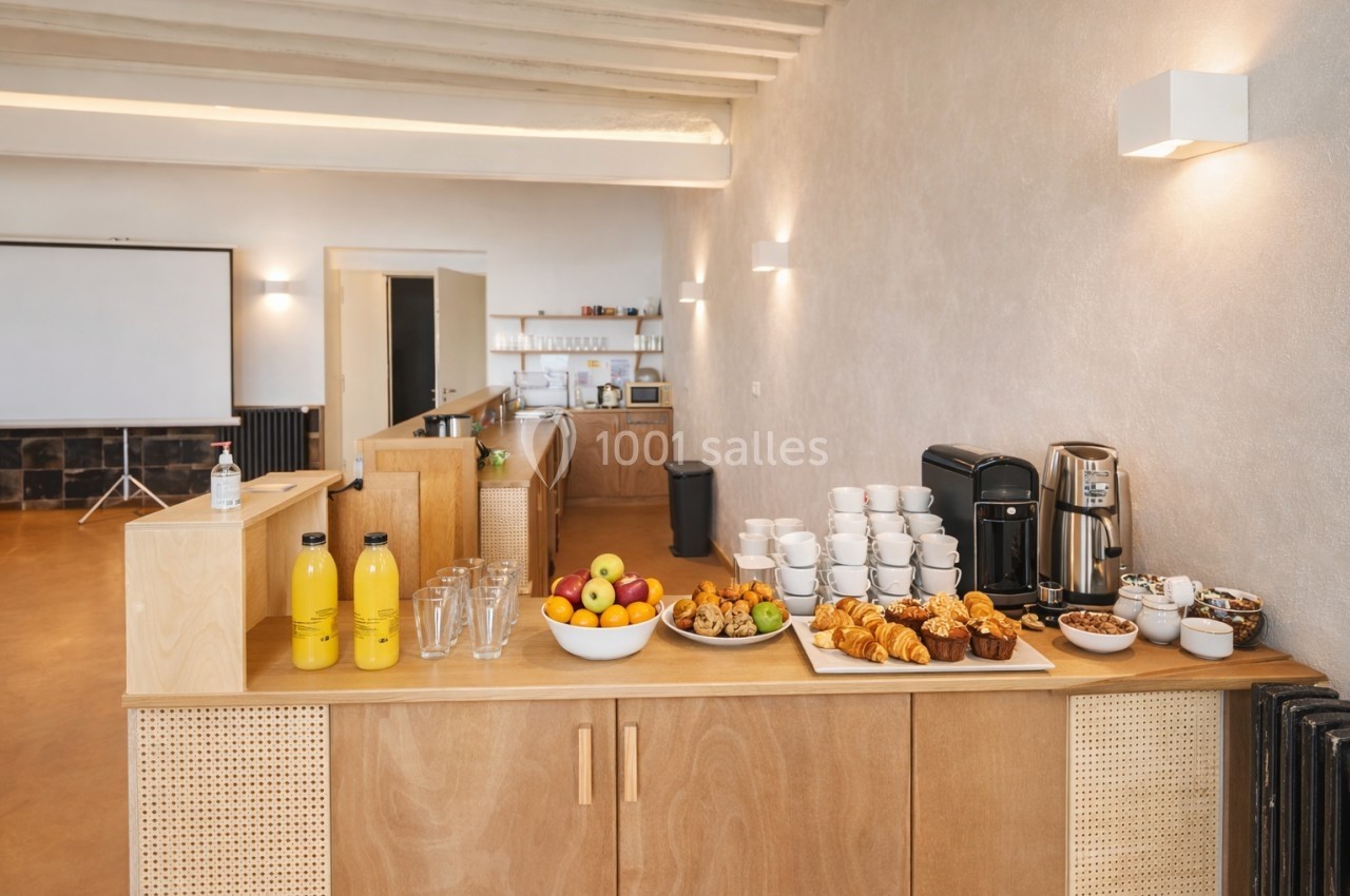 Buffet de petit-déjeuner avec viennoiseries, fruits, boissons et tasses disposés sur un comptoir en bois dans une salle…