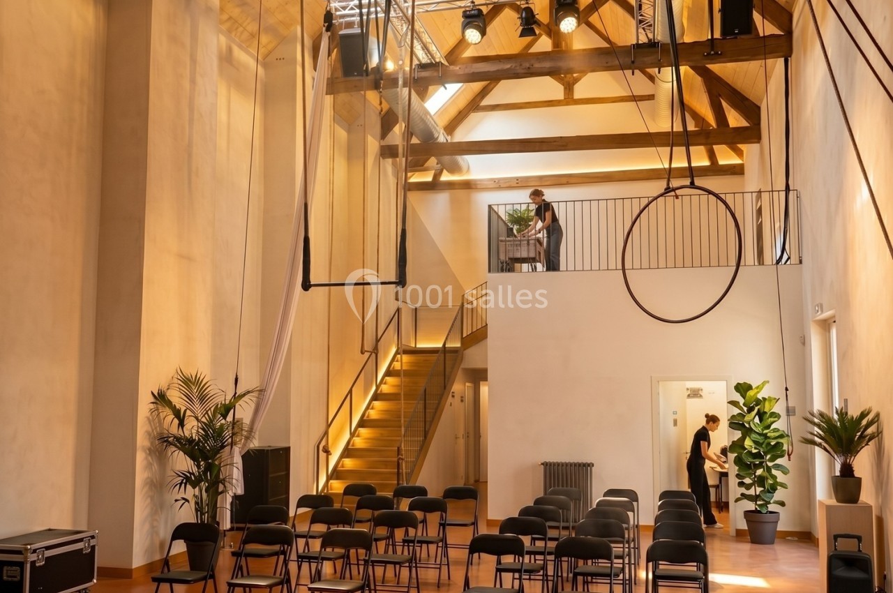 Salle lumineuse avec poutres apparentes, chaises alignées, scène surélevée et équipements techniques installés.
