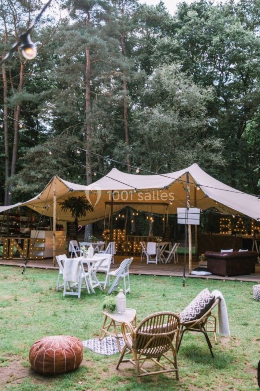 Tente beige installée en pleine forêt, avec tables, chaises et décoration pour un événement en plein air.