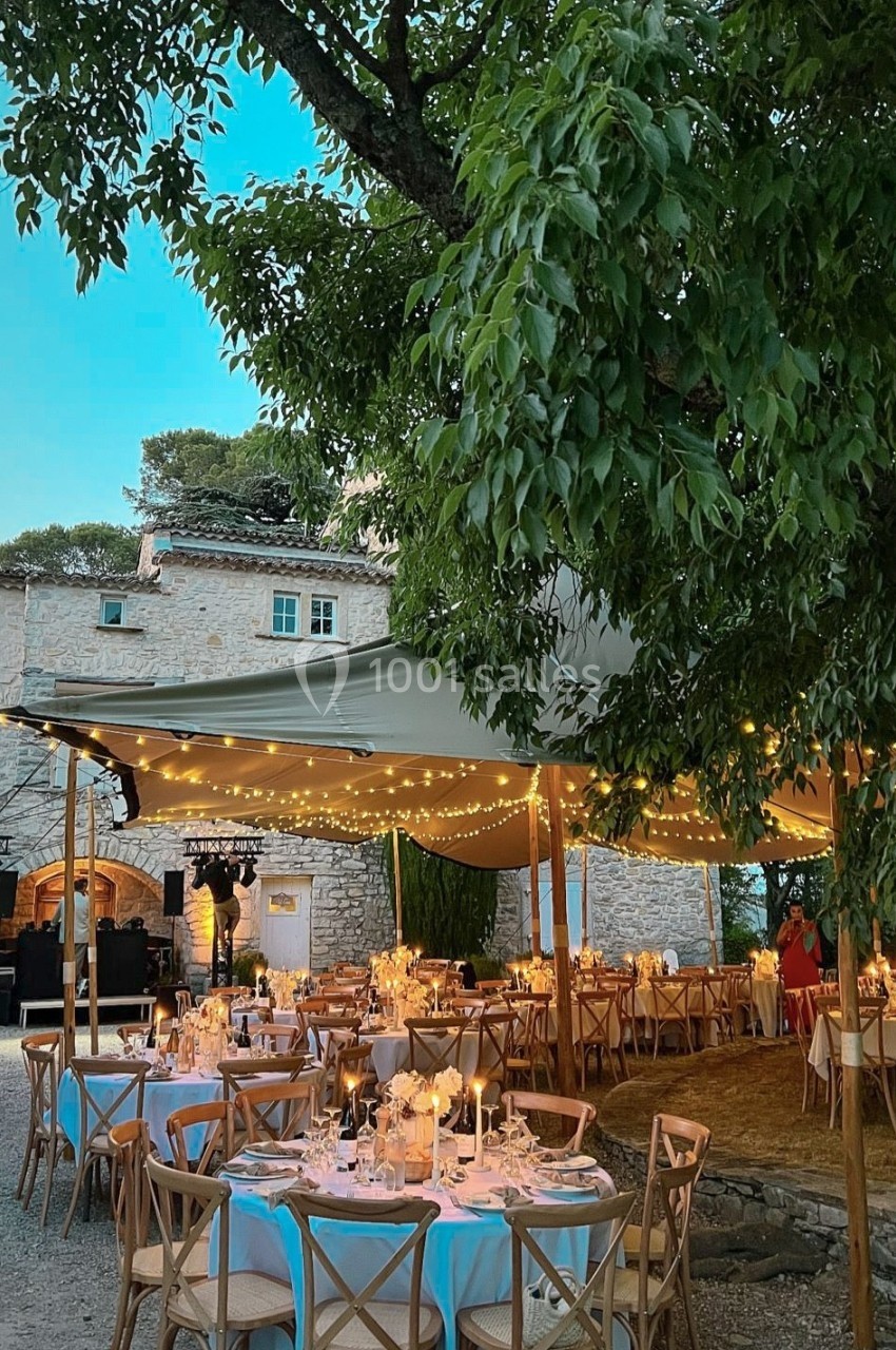 Tables élégamment dressées sous une tente éclairée par des guirlandes lumineuses, dans un jardin près d'une maison en pierre.
