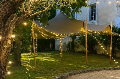 Miniature Comtralala #3 Tables décorées pour un dîner en plein air sous une tente éclairée de guirlandes lumineuses, près d'un bâtiment en pierre.