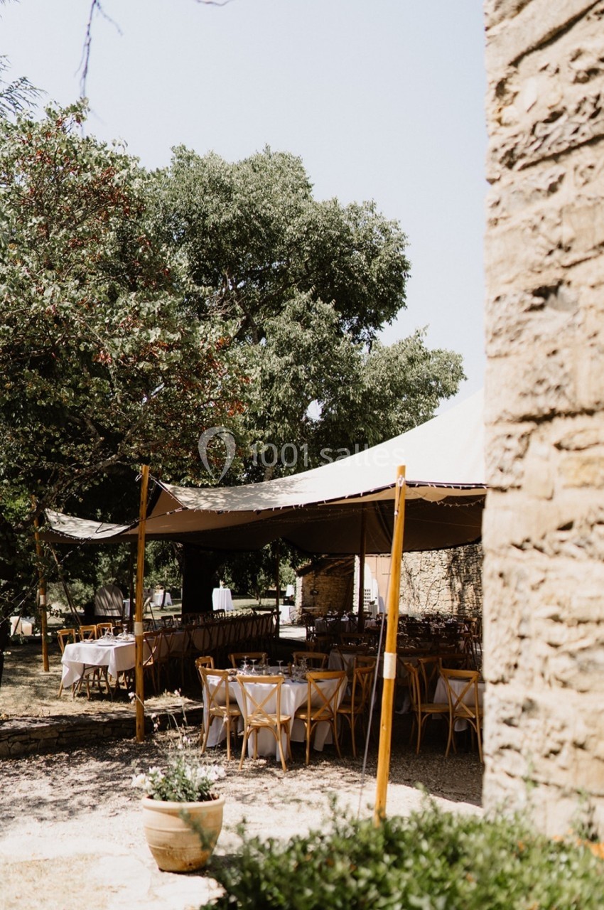 Tables dressées sous une tente en plein air, entourées d'arbres et d'un mur en pierre.