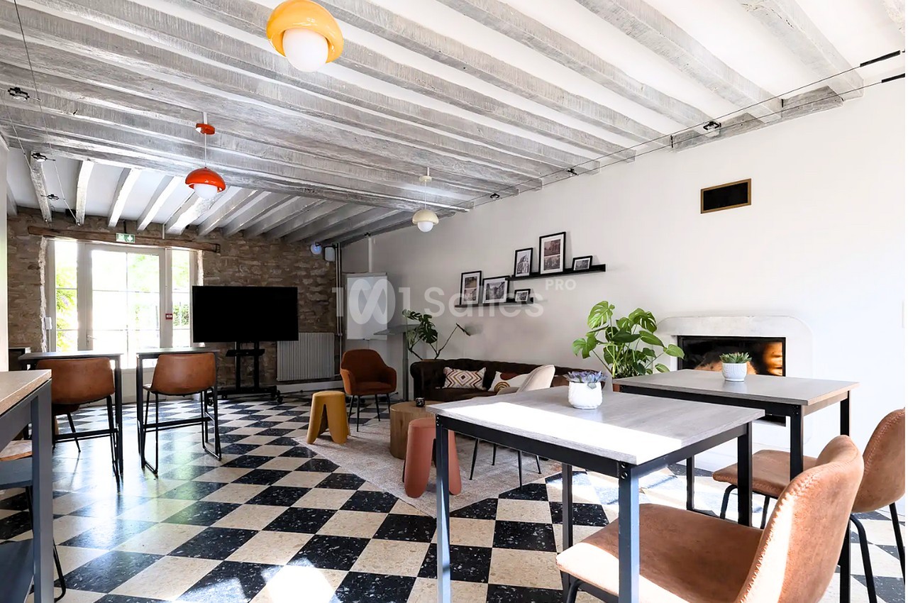 Salle à poutres apparentes aux plafonds clairs, avec tables hautes et mange-debout, canapé en cuir, cheminée et sol damier noir et blanc.