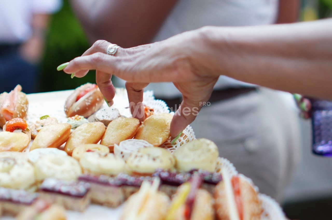 Une main féminine saisit une petite bouchée sur un plateau de mignardises salées et sucrées, en extérieur.