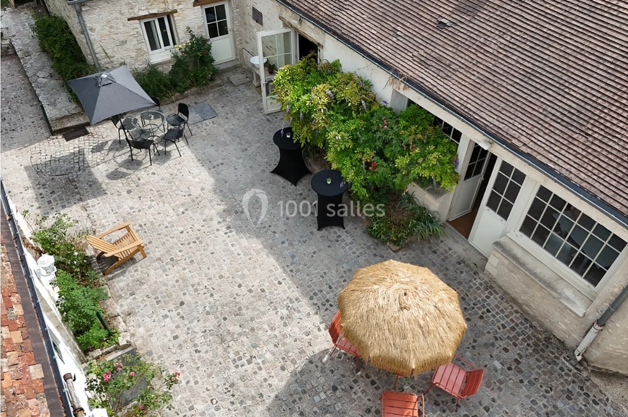 Cour pavée en vue aérienne avec mobilier de terrasse : tables hautes noires, chaises en métal, banc en bois, et parasol en paille au premier plan. Let me verify length: roughly 150 characters, too long. Need ≤125. Cour pavée vue du dessus, avec tables