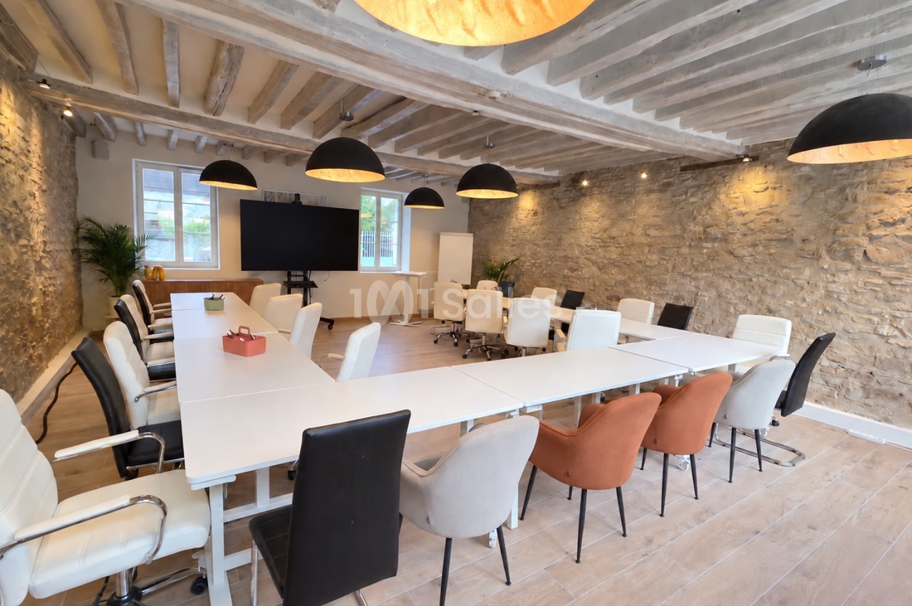 Salle de réunion aménagée en U avec de grandes tables blanches et chaises variées, murs en pierre apparente, poutres au plafond et suspensions noires. Let me check length: roughly 150 characters, too long. Need ≤125. Salle de réunion en configuration U