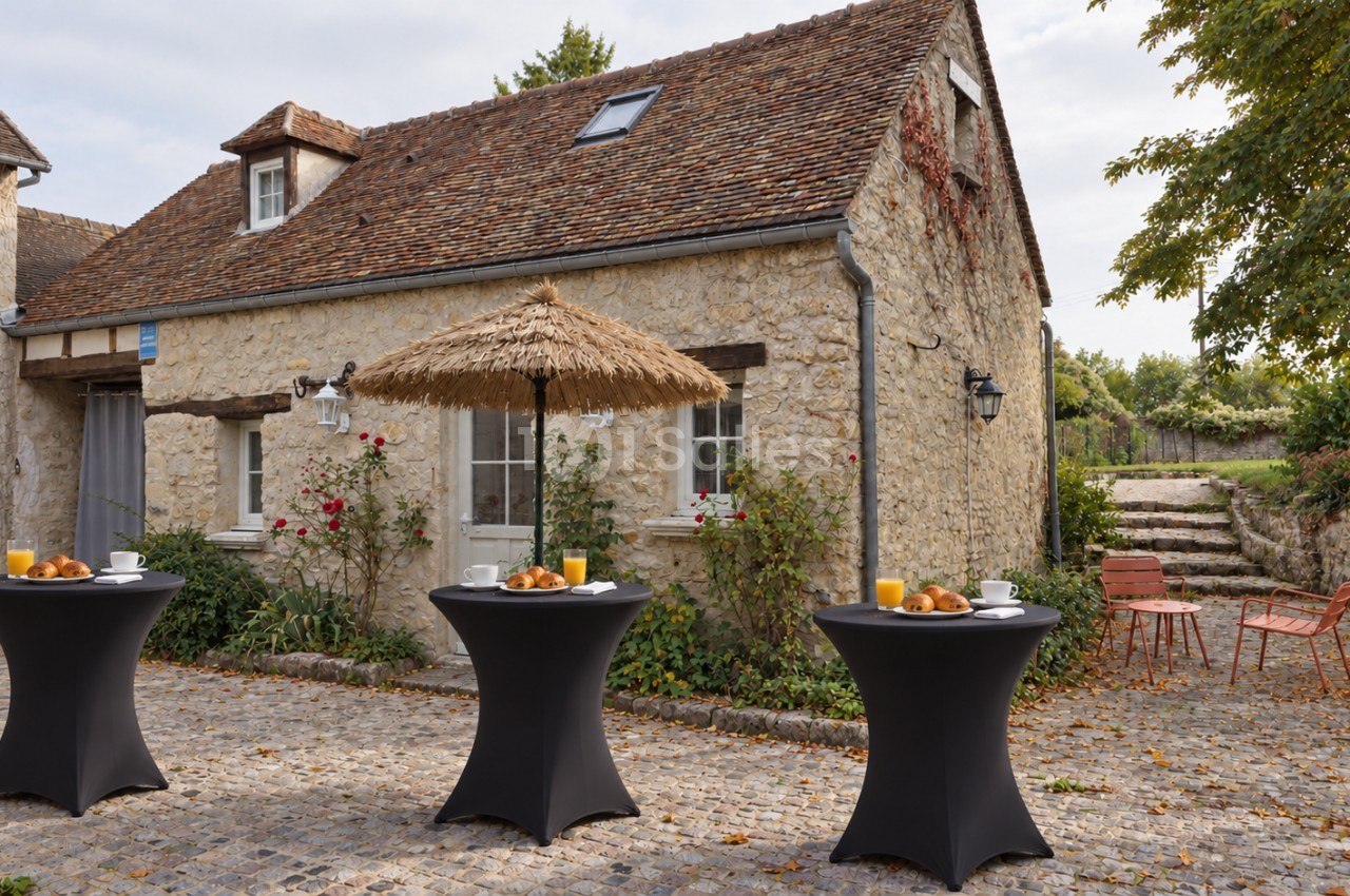Maison de pierre à toit de tuiles avec une terrasse pavée où trois tables hautes noires sont dressées de viennoiseries, jus d'orange et cafés, sous un parasol en paille. Let me check length: roughly 165 characters, too long. Need ≤125. Shorter: "Terrasse
