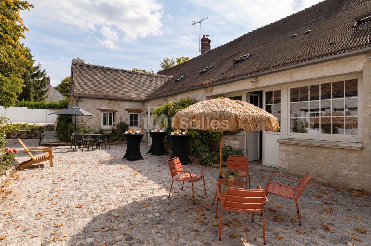 Cour pavée d'une ancienne ferme en pierre, avec tables hautes noires, mobilier orange et parasol en paille sous un ciel dégagé.