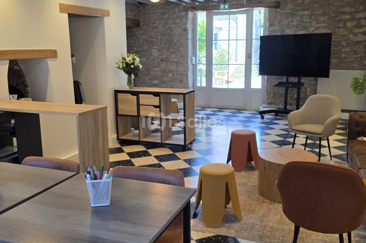 Salon aux murs en pierre apparente et sol en damier noir et blanc, avec tables de travail, fauteuils, tabourets colorés et téléviseur fixé sur pied.