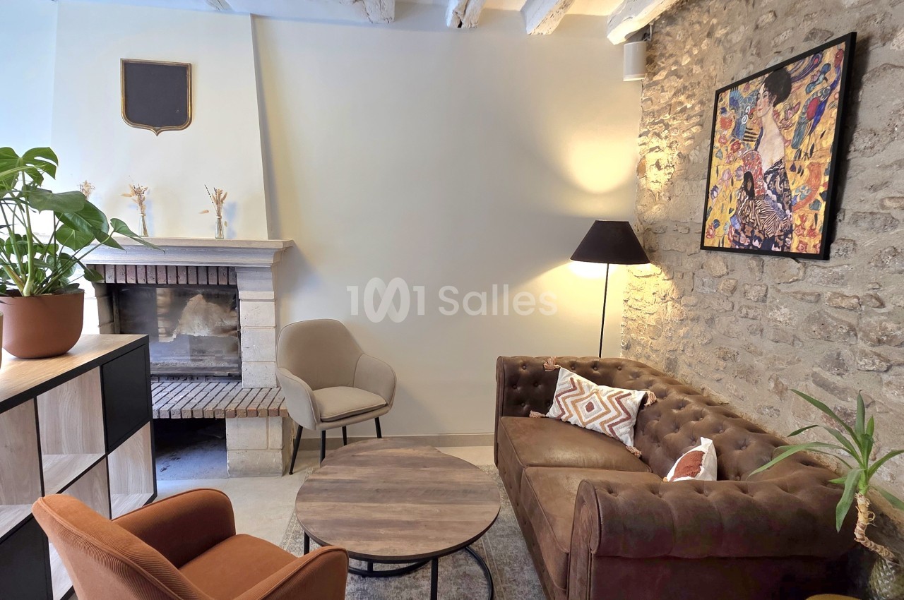 Salon au décor de pierre apparente et de poutres, avec canapé Chesterfield en cuir brun, fauteuils, table basse ronde et cheminée en pierre. Let me check length: roughly 150 characters, too long. Need ≤125. Salon aux murs de pierre apparente avec canapé