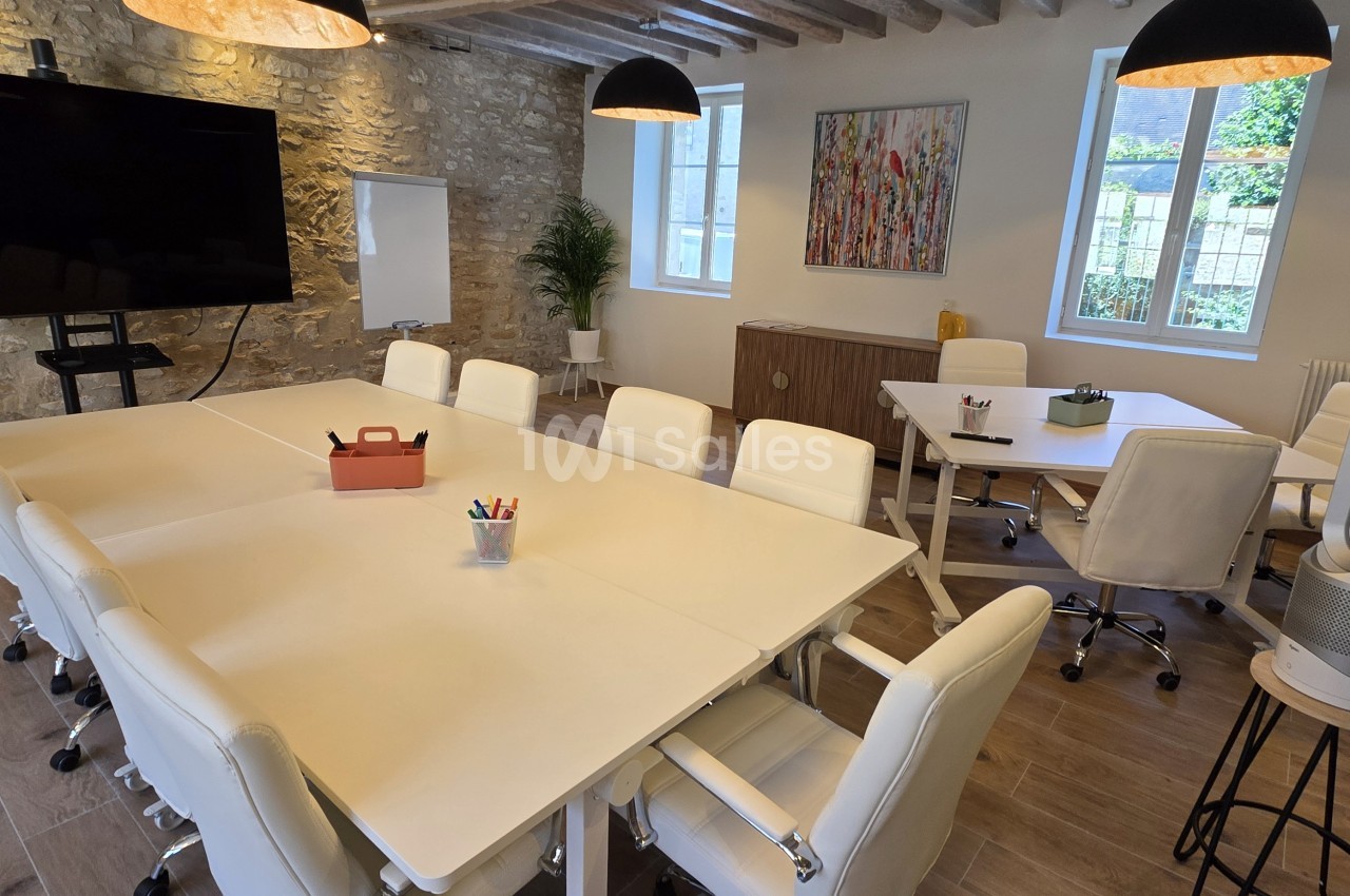 Salle de réunion aménagée de tables blanches disposées en grand rectangle, entourées de chaises blanches, avec un écran mural et un mur en pierre apparente.