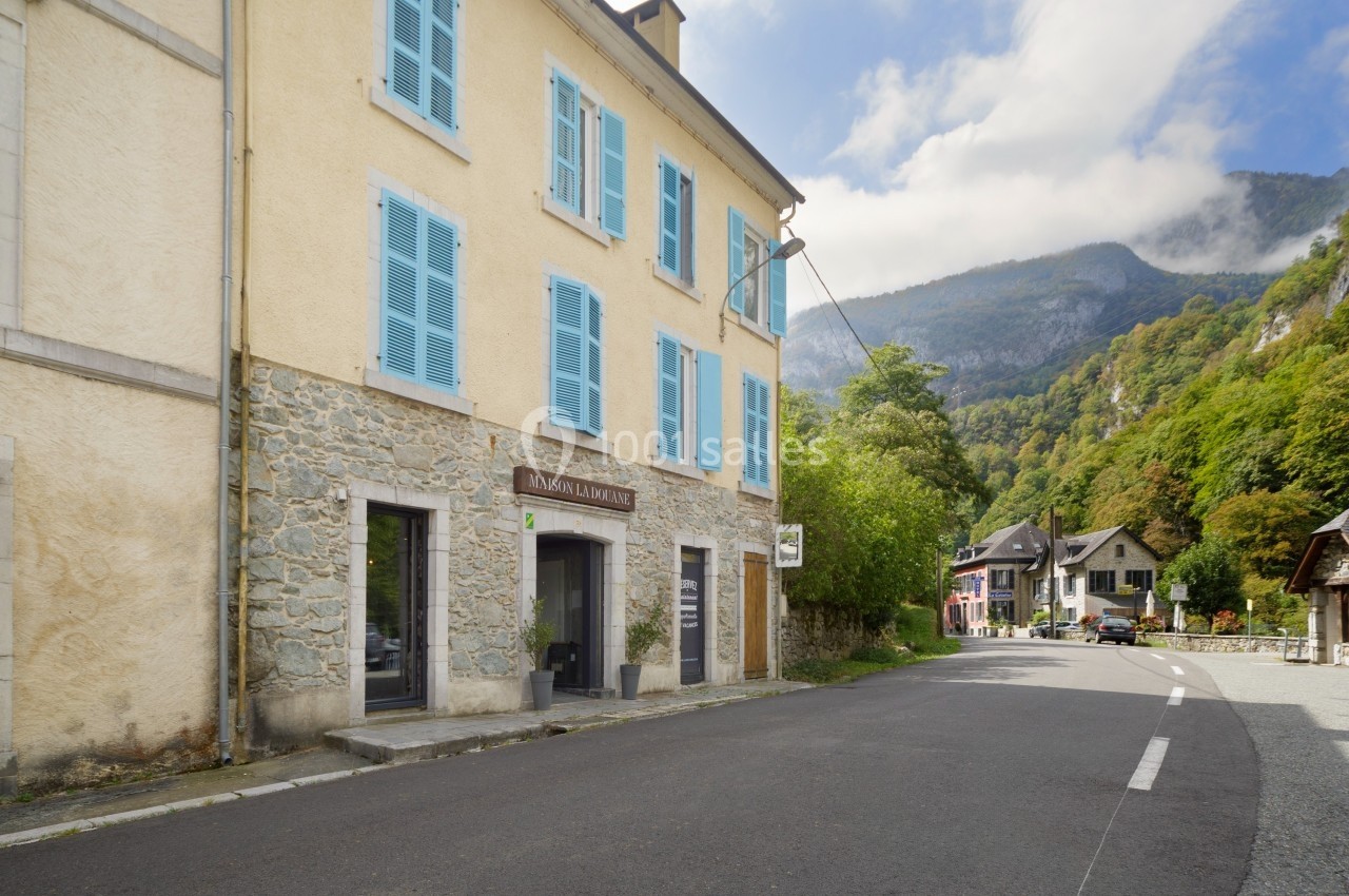 Façade d'un bâtiment en pierre avec volets bleus, situé dans un village entouré de montagnes et de végétation.