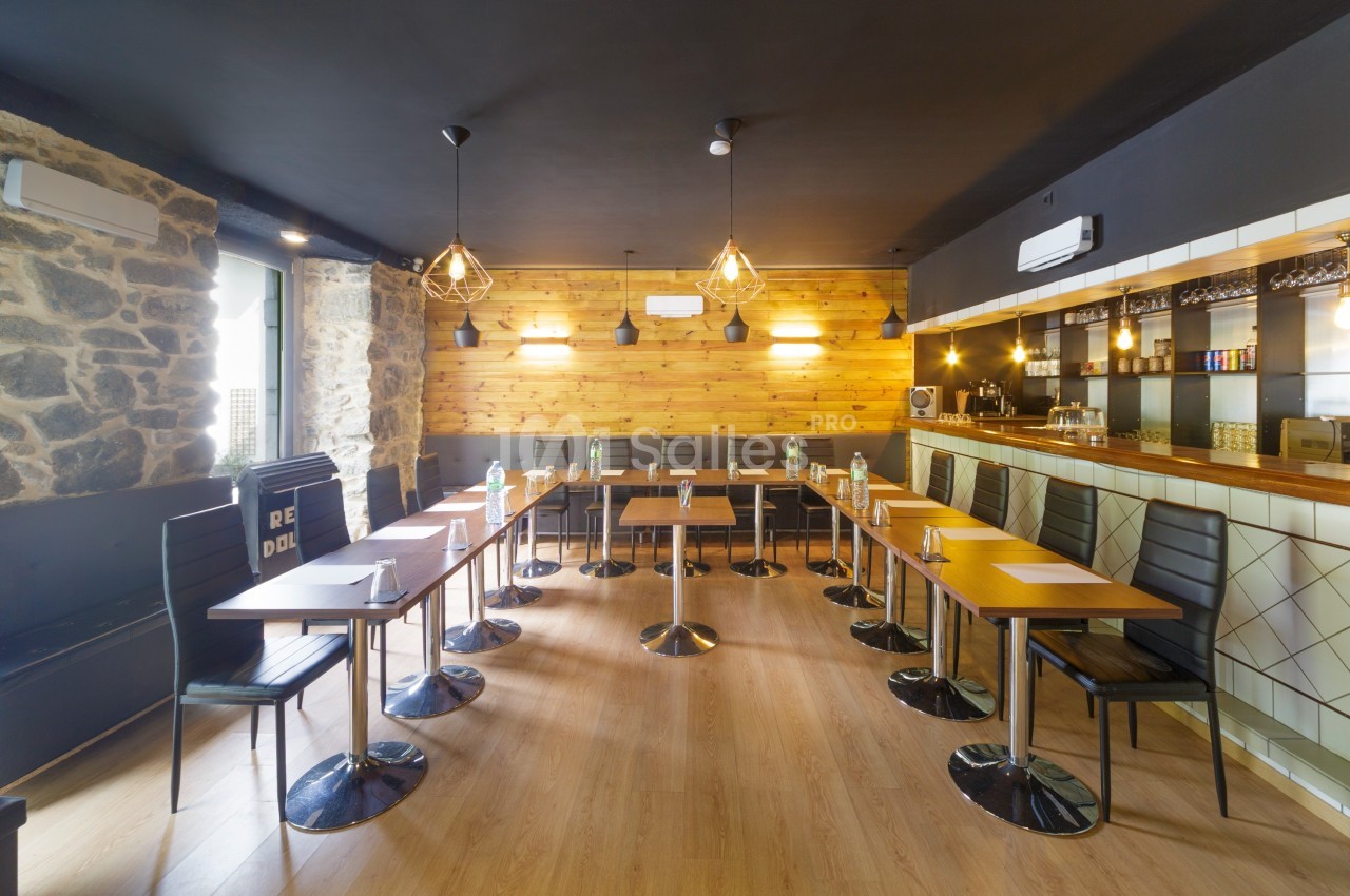 Salle de réunion lumineuse avec tables alignées, chaises noires, murs en pierre et bois, et un bar en arrière-plan.