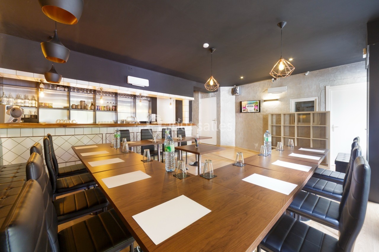 Salle de réunion moderne avec grande table en bois, chaises noires, éclairage suspendu et bar en arrière-plan.