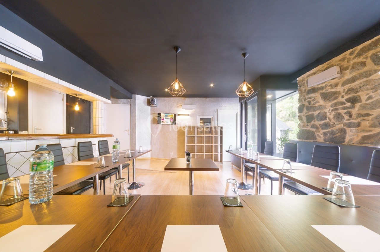 Salle de réunion lumineuse avec tables en bois, chaises noires, bouteilles d'eau et écran mural.