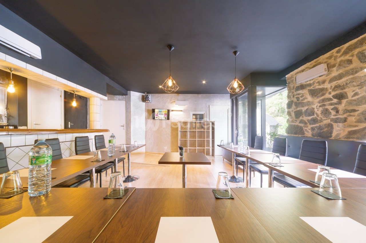 Salle de réunion lumineuse avec tables en bois, chaises noires, bouteilles d'eau et écran mural.