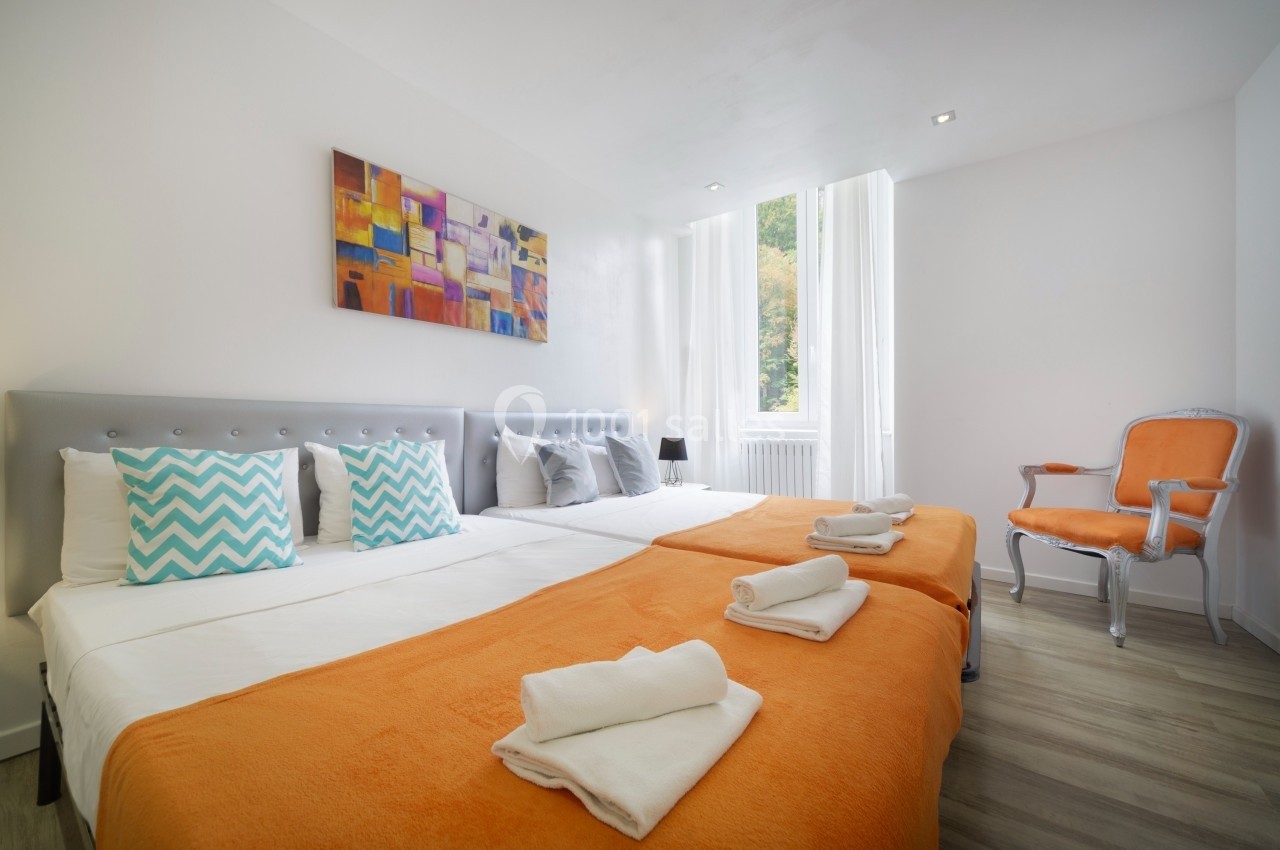 Chambre lumineuse avec deux lits simples rapprochés, draps orange, fauteuil et tableau coloré au mur.