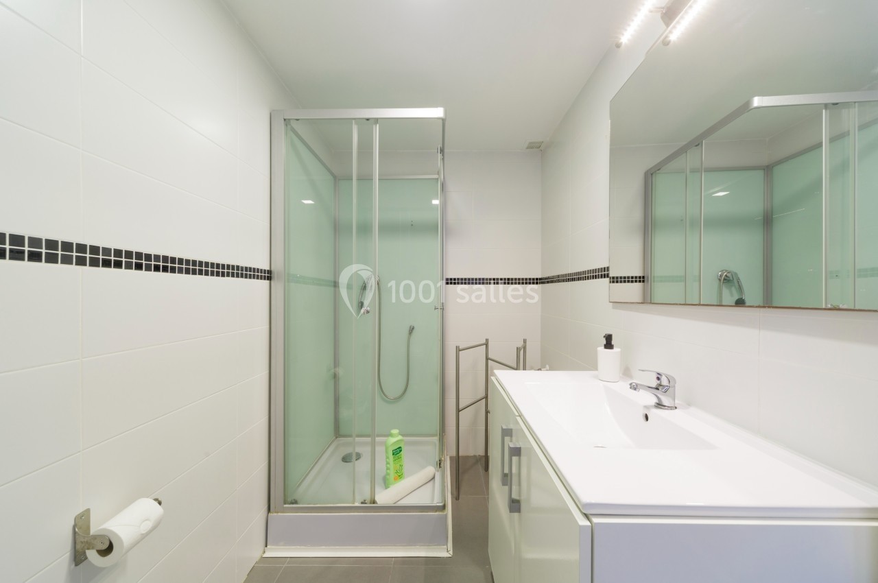 Salle de bain moderne avec douche vitrée, lavabo blanc, miroir et murs carrelés blancs avec une bande noire.