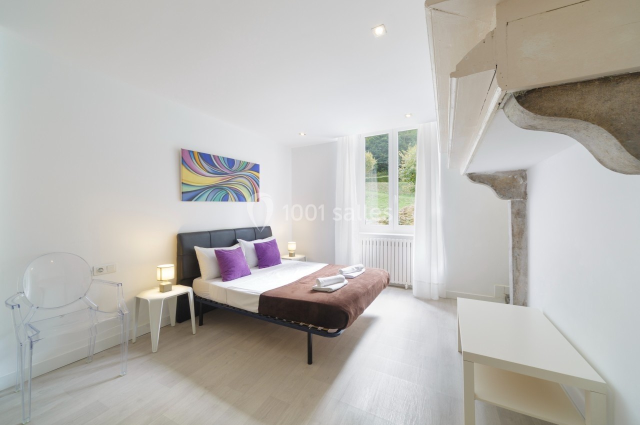 Chambre lumineuse avec lit double, coussins violets, tableau coloré au mur et grande fenêtre donnant sur un espace vert.