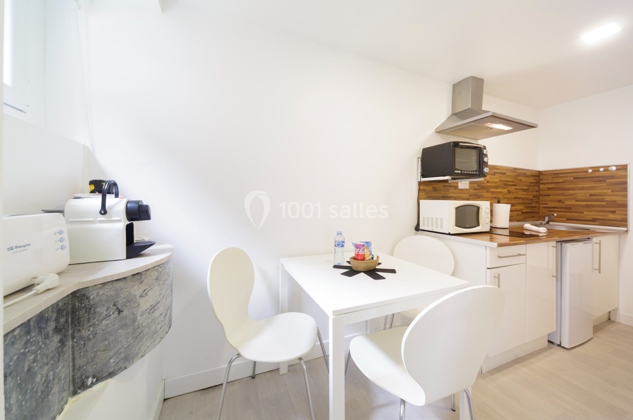 Cuisine moderne avec table blanche, trois chaises, plan de travail en bois, micro-ondes, cafetière et réfrigérateur.